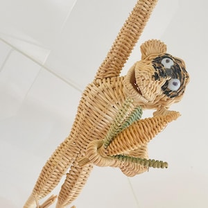May include: A hanging monkey figurine crafted from light tan woven wicker. The monkey features googly eyes, black painted facial details, and a small green woven accent on its chest. It is depicted in a suspended pose with one arm raised upward.