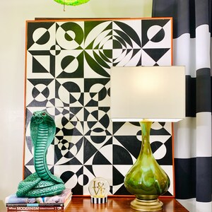 May include: A green ceramic table lamp with a white shade sits on a wooden table next to a green cobra statue and a glass globe with the word "LOVE" on it. The lamp is in front of a black and white geometric art print.