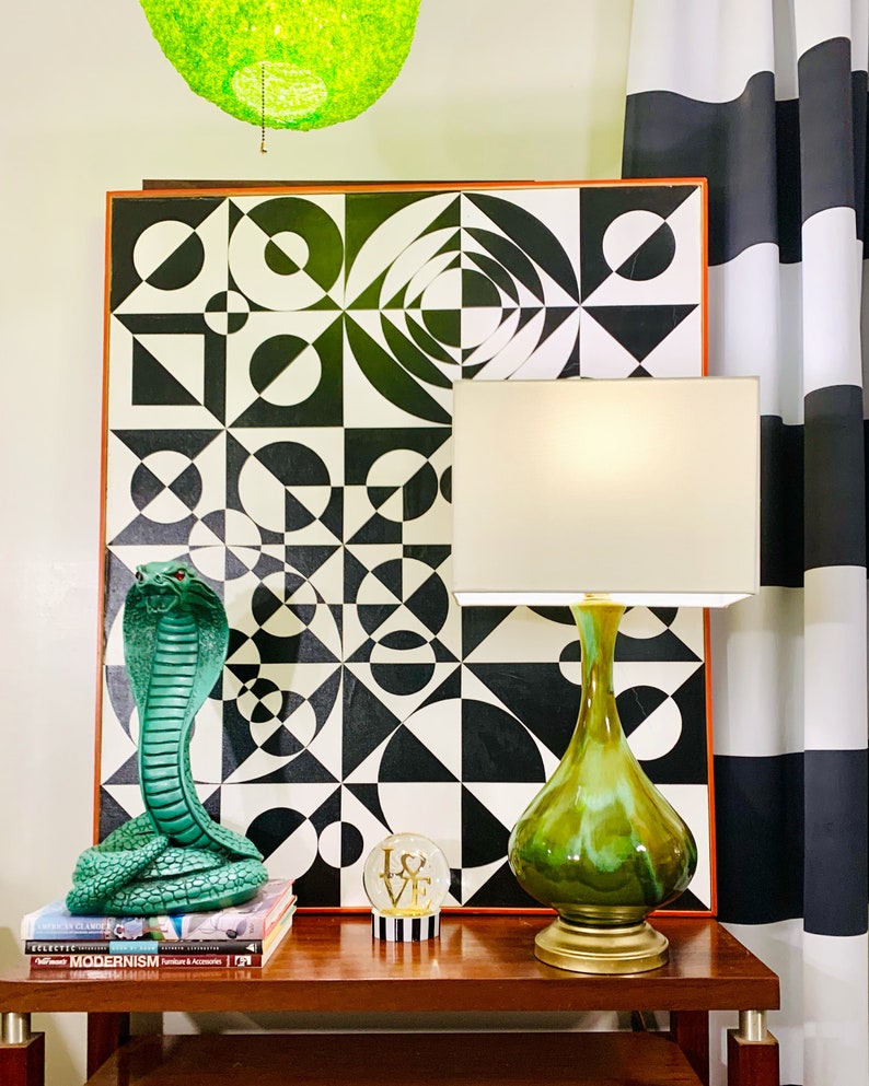 May include: A green ceramic table lamp with a white shade sits on a wooden table next to a green cobra statue and a glass globe with the word "LOVE" on it. The lamp is in front of a black and white geometric art print.