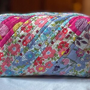 Może przedstawiać: Pikowana kosmetyczka z kwiatowym wzorem w odcieniach r&oacute;żu, błękitu i bieli. Torba ma ukośny wz&oacute;r w paski i jest prawdopodobnie ręcznie robiona. Torba jest prostokątna i ma zapięcie na zamek błyskawiczny.