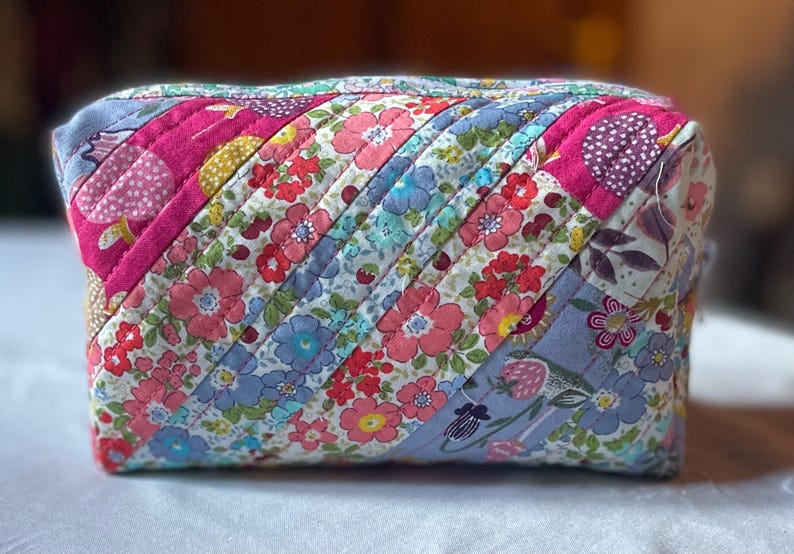 Może przedstawiać: Pikowana kosmetyczka z kwiatowym wzorem w odcieniach r&oacute;żu, błękitu i bieli. Torba ma ukośny wz&oacute;r w paski i jest prawdopodobnie ręcznie robiona. Torba jest prostokątna i ma zapięcie na zamek błyskawiczny.