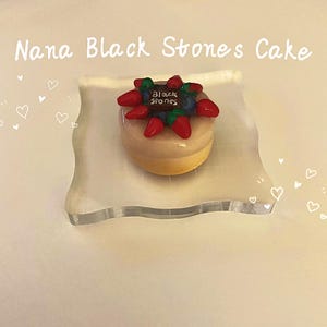 May include: A small, round cake with a light brown glaze, topped with red and blue decorations. The cake sits on a clear, wavy-edged plate. The text "Nana Black Stones Cake" is written above the cake in white script.