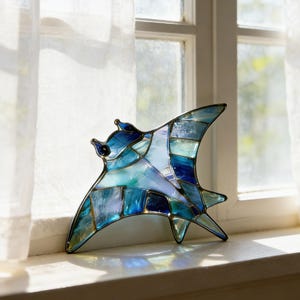 May include: A stained glass sculpture of a manta ray, featuring shades of blue, teal, and white. The artwork is composed of geometric shapes and is set against a window with sheer white curtains.