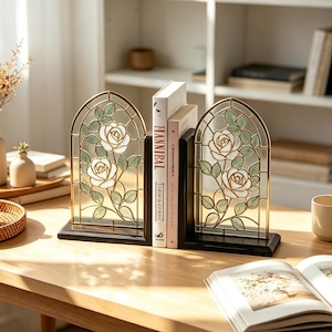 Puede incluir: Un par de sujetalibros arqueados con un diseño de vidriera de rosas blancas y hojas verdes en un marco de metal dorado sobre una base de madera negra. Los sujetalibros están colocados sobre un escritorio de madera, sosteniendo dos libros.