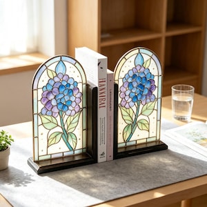May include: A pair of arched stained glass bookends featuring a blue and purple hydrangea flower design with green leaves. The bookends are set in dark wooden frames and are positioned on a desk holding two books, one titled Hannibal.