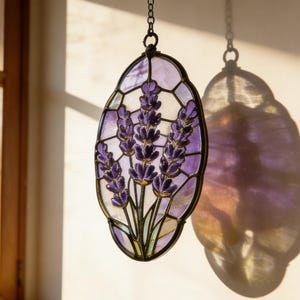 Peut inclure: Un panneau ovale en vitrail représentant des fleurs de lavande violettes et vertes. Le panneau est encadré d'une bordure en métal foncé et suspendu par une chaîne. La lumière du soleil projette une ombre sur le mur.