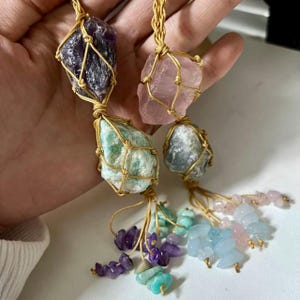 May include: Four raw gemstone pendants. The pendants feature amethyst, rose quartz, amazonite, and aquamarine stones, each encased in a woven gold-coloured cord. Small gemstone beads in various colours dangle from the bottom of each pendant.