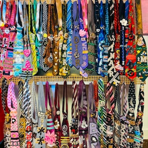 May include: A collection of neckties displayed on a gold grid, featuring various colors, patterns, and embellishments including pins, patches, fabric flowers, and lettered text. The ties are arranged in two rows, showcasing diverse designs and textures.