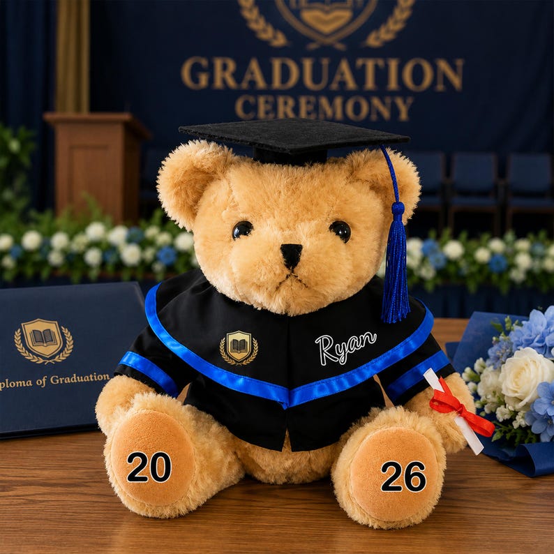 Puede incluir: Un oso de peluche con atuendo de graduaci&oacute;n. El oso lleva birrete y toga negros con ribetes azules. El nombre "Ryan" est&aacute; en la toga. Las patas del oso muestran los n&uacute;meros "20" y "26". Tambi&eacute;n se ven un diploma y flores.