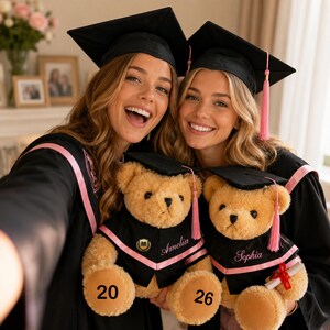 Puede incluir: Dos graduadas sonriendo, vestidas con togas y birretes negros con detalles rosas, sosteniendo osos de peluche vestidos con atuendos de graduaci&oacute;n en miniatura. Los osos tienen los nombres "Amelia" y "Sophia", y los n&uacute;meros "20" y "26".