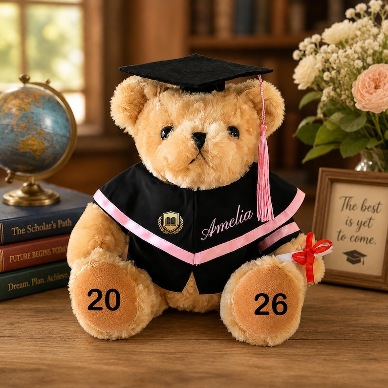 Puede incluir: Un oso de peluche con un atuendo de graduaci&oacute;n, que incluye un gorro y una toga negros con ribetes rosas. El nombre "Amelia" est&aacute; impreso en la toga. El oso tiene los n&uacute;meros "20" y "26" en sus patas y sostiene un diploma con una cinta roja.