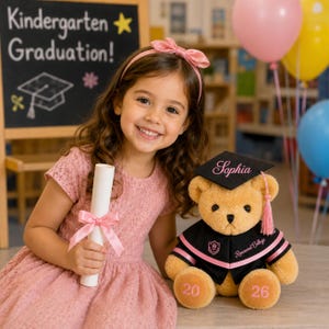 Puede incluir: Una ni&ntilde;a con un vestido y lazo rosa sonr&iacute;e, sosteniendo un diploma atado con una cinta rosa. A su lado, un oso de peluche lleva un birrete con el nombre "Sophia" y una toga negra con ribete rosa. La pizarra dice "Kindergarten Graduation!"