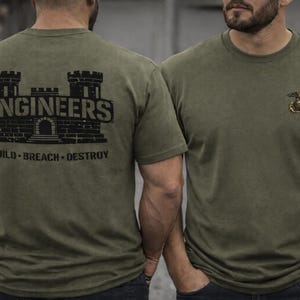 May include: Olive green t-shirts. One shirt features a black graphic of a castle with the word "ENGINEERS" above and "BUILD - BREACH - DESTROY" below. The other shirt has a small emblem on the chest.