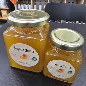 May include: Two glass jars of mango jam with gold lids. The jars have labels that read "Joyous Jams" and "Mango Jam". The jam is a bright yellow color. The jars are on a dark surface.