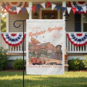 May include: A white garden flag featuring a colorful illustration of Radiator Springs, with the text "Welcome to Radiator Springs." The flag includes images of a red race car, desert landscapes, and the text "California Est. 2017."