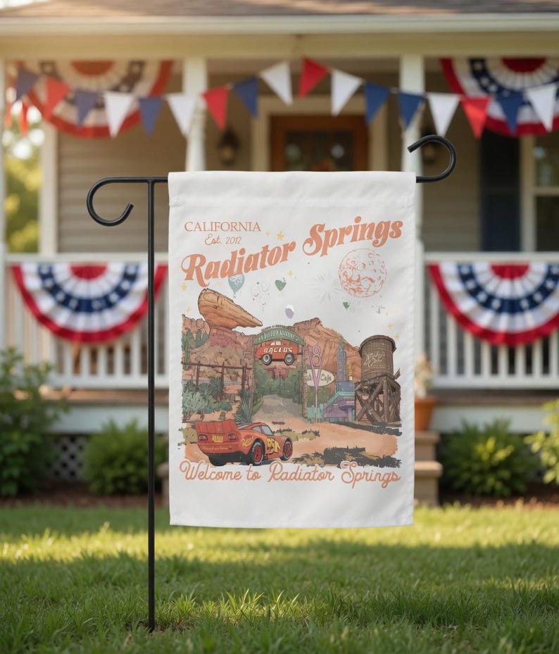 May include: A white garden flag featuring a colorful illustration of Radiator Springs, with the text "Welcome to Radiator Springs." The flag includes images of a red race car, desert landscapes, and the text "California Est. 2017."