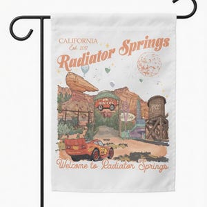 May include: A white garden flag featuring a colorful illustration of Radiator Springs, with the text "CALIFORNIA Est. 2012 Radiator Springs" and "Welcome to Radiator Springs." The design includes a red race car and desert landscape.