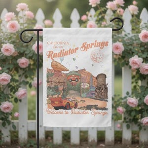 May include: A white garden flag featuring a colorful illustration of Radiator Springs, with text that reads "CALIFORNIA Est. 2012 Radiator Springs" and "Welcome to Radiator Springs." The design includes a red race car, desert scenery, and a water tower.