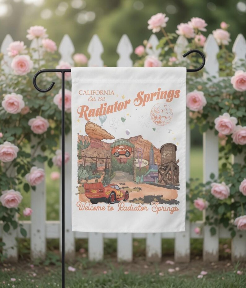 May include: A white garden flag featuring a colorful illustration of Radiator Springs, with text that reads "CALIFORNIA Est. 2012 Radiator Springs" and "Welcome to Radiator Springs." The design includes a red race car, desert scenery, and a water tower.