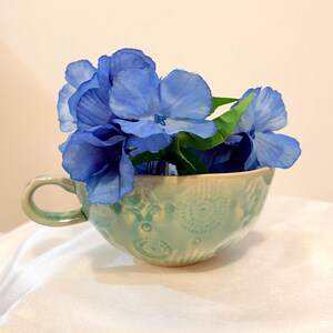 Peut inclure: Une tasse à thé en céramique turquoise remplie de fleurs bleues. La tasse présente une anse et un motif en relief. Les fleurs bleues contrastent avec la couleur de la tasse, créant un arrangement visuellement attrayant.