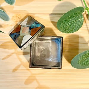 May include: A silver-toned square box with an open lid, revealing the interior. The lid is decorated with a geometric pattern of inlaid wood, abalone shell, and other materials in brown, blue, and white. The box rests on a wooden surface.