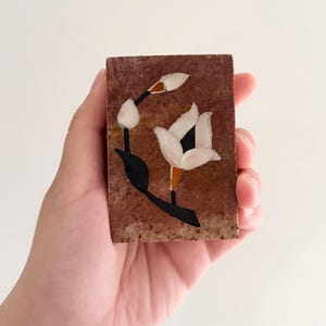 Puede incluir: Una placa rectangular de piedra con una superficie de color marrón rojizo moteado que presenta un diseño floral incrustado. La incrustación consta de pétalos blancos, tallos y hojas negras, y pequeños acentos naranjas sobre la piedra.