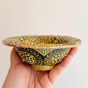 May include: A small, round ceramic bowl featuring a vibrant, hand-painted pattern of dark green, yellow, and orange geometric shapes. The exterior and wide rim are decorated with intricate, raised dot details in white, blue, and orange, creating a textured surface.