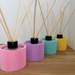 May include: Four cylindrical reed diffusers with ribbed exterior shells in pastel pink, lavender, light blue, and yellow. Each diffuser features a black circular cap at the centre and several thin, light-coloured wooden reeds extending upwards.