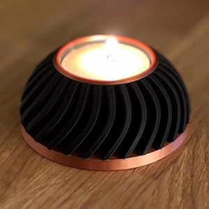 May include: A black and copper candle holder with a lit candle. The holder features a spiral design and a copper rim. The candle flame glows brightly, casting a warm light. The candle holder is a decorative item.