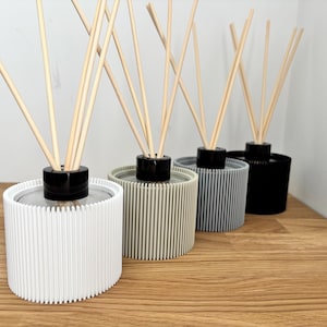 Puede incluir: Cuatro difusores de varillas con recipientes cilíndricos de estrías verticales en blanco, beige, gris y negro. Cada difusor cuenta con una tapa circular negra en el centro y varias varillas de madera clara que sobresalen hacia arriba.