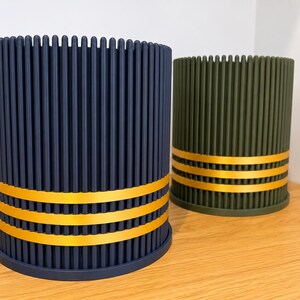 Peut inclure: Deux récipients cylindriques avec des textures nervurées verticales. L'un est bleu marine foncé et l'autre est vert olive. Les deux présentent trois bandes dorées horizontales enroulées autour de la partie inférieure, créant un contraste.