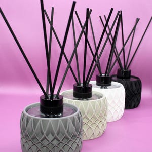 May include: Four reed diffusers arranged in a row, each featuring a cylindrical container with a geometric lattice pattern. The containers are coloured grey, beige, white, and black, topped with black caps and several thin black diffuser reeds.
