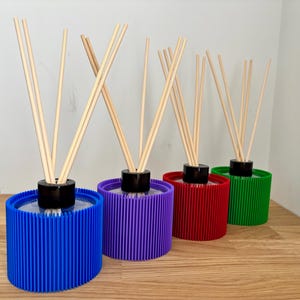 May include: Four cylindrical reed diffusers with ribbed exterior walls in blue, purple, red, and green. Each diffuser features a black circular cap at the centre with several light-coloured wooden reeds extending upwards from the opening.