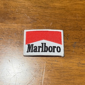 May include: A rectangular embroidered patch featuring a white background with a red upper section and the black text Marlboro centred in the lower portion. The patch has a white stitched border and a flat, fabric-based construction.