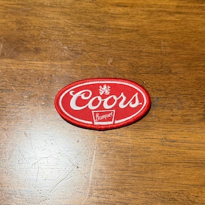May include: An oval-shaped embroidered patch featuring a red background with a white border. The text Coors is stitched in white script, accompanied by a small white emblem above and the word Banquet in a white rectangular outline below.