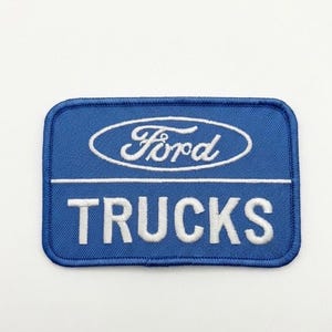 May include: A rectangular embroidered patch with a blue background and a dark blue border. The patch features the white Ford logo inside an oval at the top, a horizontal white line, and the word TRUCKS in white capital letters below.
