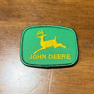 May include: A rectangular embroidered patch with rounded corners featuring a bright green background and a yellow leaping deer silhouette. The text JOHN DEERE is embroidered in yellow capital letters below the deer, all bordered by green stitching.