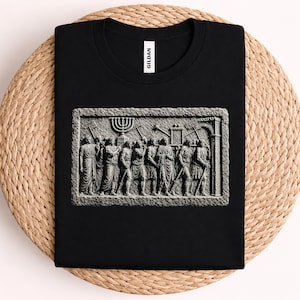 May include: A black crew neck t-shirt featuring a rectangular graphic print of a stone relief carving. The print depicts a group of figures in ancient-style robes carrying a menorah and various tools, with the brand name GILDAN on the neck label.