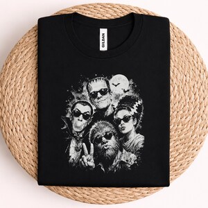 May include: A black short-sleeve t-shirt featuring a white graphic print of four classic horror characters wearing sunglasses. The design includes a full moon, bats, and the characters posing together. The shirt has a Gildan brand neck label.