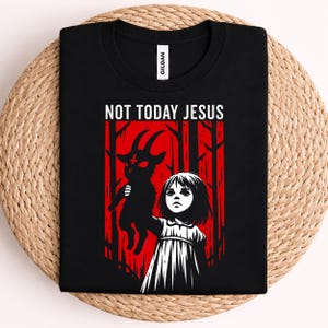 May include: A black t-shirt featuring a graphic print of a person holding a black goat against a red forest background. The text "NOT TODAY JESUS" is printed in white above the illustration. The shirt has a white Gildan brand neck label.