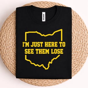 May include: A black short-sleeved t-shirt featuring a yellow outline of the state of Ohio. Centered within the state outline is the yellow text "I'M JUST HERE TO SEE THEM LOSE" in a bold, blocky font. The shirt has a Gildan brand neck label.