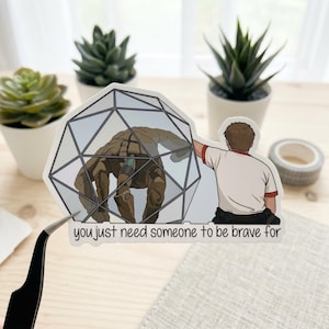 May include: A sticker featuring a brown creature inside a geometric cage, with a person reaching out to it. The sticker has the text "you just need someone to be brave for". The background includes small potted plants and a roll of tape.