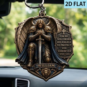 Könnte beinhalten: Ein flaches 2D-Hängeornament mit einem knienden Engelskrieger in bronzefarbener Rüstung mit großen Flügeln, der ein Schwert hält. Eine Plakette rechts trägt die Inschrift FOR HE WILL ORDER HIS ANGELS TO PROTECT YOU WHEREVER YOU GO. Unten steht WILLIAM.