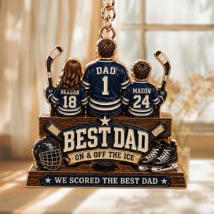 May include: A hockey-themed ornament featuring a father and two children in hockey jerseys. The ornament reads "BEST DAD" and includes a hockey helmet, puck, and skates. The jerseys display the names "DAD", "Reagan", and "Mason" with numbers.
