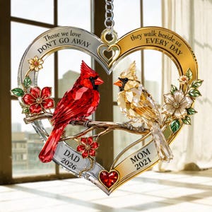 Op de afbeelding: Een hartvormig hangend ornament met een rode kardinaal en een goudkleurige vogel op een tak. Het frame bevat de tekst "Those we love DON'T GO AWAY" en "they walk beside us EVERY DAY" met de inscripties "DAD 2026" en "MOM 2021".