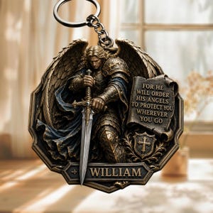 May include: A circular metal keychain featuring a relief of a winged warrior in armor holding a sword. A scroll displays the text: FOR HE WILL ORDER HIS ANGELS TO PROTECT YOU WHEREVER YOU GO. Below, a shield and a nameplate with the name WILLIAM.