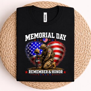 May include: Black t-shirt with a Memorial Day graphic. The design features a heart-shaped American flag, a kneeling soldier, and the words "MEMORIAL DAY" and "REMEMBER & HONOR" in white. The shirt is on a woven surface.
