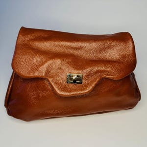 Puede incluir: Un bolso de mano de cuero marrón con cierre de solapa y cierre dorado. El bolso tiene una superficie texturizada y una forma rectangular. La solapa se pliega sobre la parte superior del bolso.