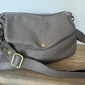 May include: A taupe leather crossbody bag featuring a fold-over flap with a single brass-toned snap closure. The bag includes an adjustable shoulder strap attached with brass-toned metal hardware, including a sliding buckle and two rivets.