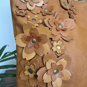 Puede incluir: Un bolso tote de cuero marrón claro con múltiples aplicaciones de flores de cuero en varios tonos de marrón. Las flores están aseguradas con remaches de color latón. El bolso tiene un asa con herrajes de color latón.